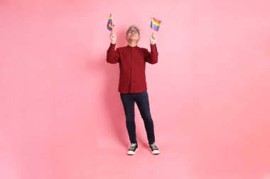 İki elinde de pembe arka planda izole edilmiş Rainbown Pride Bayrakları olan resmi ve sıradan kırmızı tişörtlü Asyalı kıdemli bir adam. LGBTQ + topluluğu