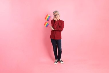 İki elinde de pembe arka planda izole edilmiş Rainbown Pride Bayrakları olan resmi ve sıradan kırmızı tişörtlü Asyalı kıdemli bir adam. LGBTQ + topluluğu