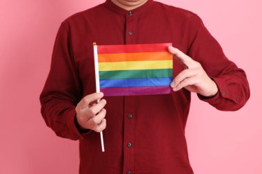 İki elinde de pembe arka planda izole edilmiş Rainbown Pride Bayrakları olan resmi ve sıradan kırmızı tişörtlü Asyalı kıdemli bir adam. LGBTQ + topluluğu