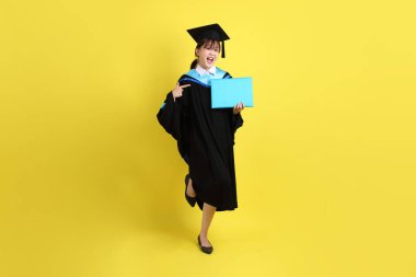 Mavi mezuniyet elbiseleri ve kepleri içinde kadın üniversite öğrencisi ve sarı arka planda izole edilmiş işaret eden bir jestle diplomaları var. Gen Z, yeni mezun, eğitim, ilk iş