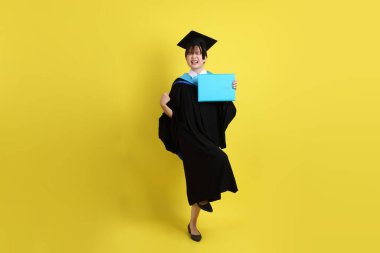 Mavi mezuniyet elbiseleri ve kepleri içinde, elinde diplomaları olan, heyecanlı, sarı arka planda izole bir şekilde kutlanan bir üniversite öğrencisi. Gen Z, yeni mezun, eğitim, ilk iş