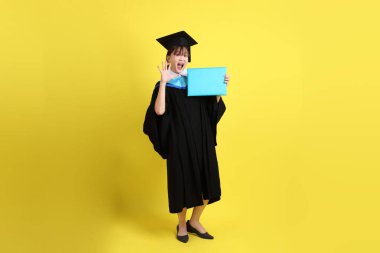 Mavi mezuniyet elbiseleri ve kepleri içinde, jest sürprizi ile diplomaları olan ve sarı arka planda izole edilmiş etkileyici bir üniversite öğrencisi. Gen Z, yeni mezun, eğitim, ilk iş