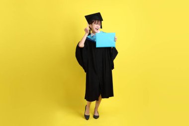 Mavi mezuniyet elbiseleri ve kepleri içinde kadın üniversite öğrencisi ve sarı arka planda izole edilmiş işaret eden bir jestle diplomaları var. Gen Z, yeni mezun, eğitim, ilk iş