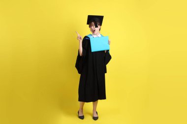 Mavi mezuniyet elbiseleri ve kepleri içinde kadın üniversite öğrencisi ve sarı arka planda izole edilmiş işaret eden bir jestle diplomaları var. Gen Z, yeni mezun, eğitim, ilk iş