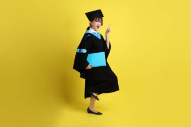 Mavi mezuniyet elbiseleri ve kepleri içinde kadın üniversite öğrencisi ve sarı arka planda izole edilmiş işaret eden bir jestle diplomaları var. Gen Z, yeni mezun, eğitim, ilk iş