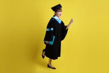 Mavi mezuniyet elbiseleri ve kepleri içinde kadın üniversite öğrencisi ve sarı arka planda izole bir şekilde yürüme jesti ile diplomaları var. Gen Z, yeni mezun, eğitim, ilk iş