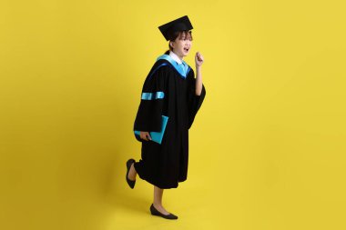 Mavi mezuniyet elbiseleri ve kepleri içinde kadın üniversite öğrencisi ve sarı arka planda izole bir şekilde yürüme jesti ile diplomaları var. Gen Z, yeni mezun, eğitim, ilk iş