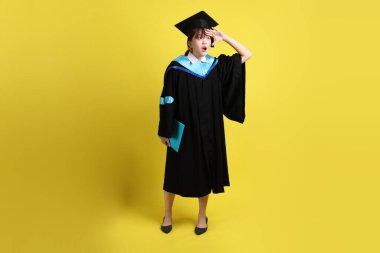 Mavi mezuniyet elbiseleri ve kepleri içinde, elinde diplomalarıyla, sarı arka planda izole edilmiş uzaklığa bakma hareketi yapan bir üniversite öğrencisi. Gen Z, yeni mezun, eğitim, ilk iş