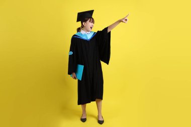 Mavi mezuniyet elbiseleri ve kepleri içinde kadın üniversite öğrencisi ve sarı arka planda izole edilmiş işaret eden bir jestle diplomaları var. Gen Z, yeni mezun, eğitim, ilk iş