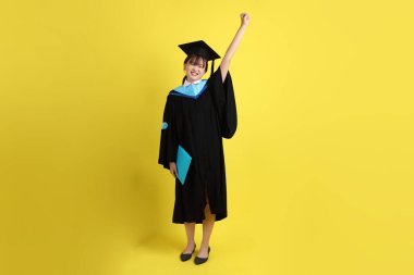 Mavi mezuniyet elbiseleri ve kepleri içinde, elinde diplomaları olan, heyecanlı, sarı arka planda izole bir şekilde kutlanan bir üniversite öğrencisi. Gen Z, yeni mezun, eğitim, ilk iş