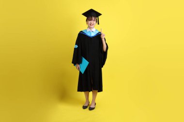 Mavi mezuniyet elbiseleri ve şapkaları içinde, elinde diplomaları olan, sarı arka planda bir numaralı jesti olan bir üniversite öğrencisi. Gen Z, yeni mezun, eğitim, ilk iş