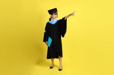 Mavi mezuniyet elbiseleri ve kepleri içinde kadın üniversite öğrencisi ve sarı arka planda izole edilmiş işaret eden bir jestle diplomaları var. Gen Z, yeni mezun, eğitim, ilk iş