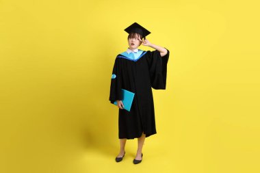 Mavi mezuniyet elbiseleri ve kepleri içinde kadın üniversite öğrencisi ve sarı arka planda izole edilmiş düşünce jestleriyle diplomaları var. Gen Z, yeni mezun, eğitim, ilk iş