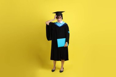 Mavi mezuniyet elbiseleri ve kepleri içinde kadın üniversite öğrencisi ve sarı arka planda izole edilmiş düşünce jestleriyle diplomaları var. Gen Z, yeni mezun, eğitim, ilk iş