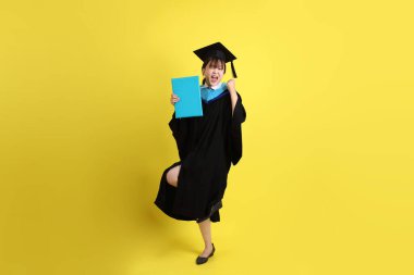 Mavi mezuniyet elbiseleri ve kepleri içinde, elinde diplomaları olan, heyecanlı, sarı arka planda izole bir şekilde kutlanan bir üniversite öğrencisi. Gen Z, yeni mezun, eğitim, ilk iş