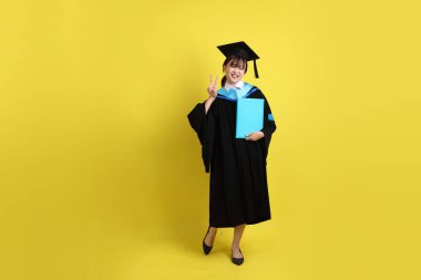 Mavi mezuniyet elbiseleri ve kepleri içinde, elinde diplomaları olan, barış işareti olan, sarı arka planda izole edilmiş bir üniversite öğrencisi. Gen Z, yeni mezun, eğitim, ilk iş