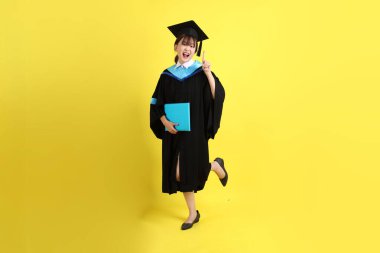 Mavi mezuniyet elbiseleri ve şapkaları içinde, elinde diplomaları olan, sarı arka planda bir numaralı jesti olan bir üniversite öğrencisi. Gen Z, yeni mezun, eğitim, ilk iş