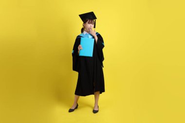 Mavi mezuniyet elbiseleri ve kepleri içinde, jest sürprizi ile diplomaları olan ve sarı arka planda izole edilmiş etkileyici bir üniversite öğrencisi. Gen Z, yeni mezun, eğitim, ilk iş