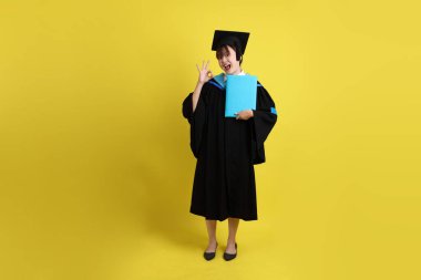 Mavi mezuniyet elbiseleri ve kepleri içinde, elinde diplomaları olan, baş parmak kaldıran ya da sarı arka planda izole edilmiş bir üniversite öğrencisi. Gen Z, yeni mezun, eğitim, ilk iş