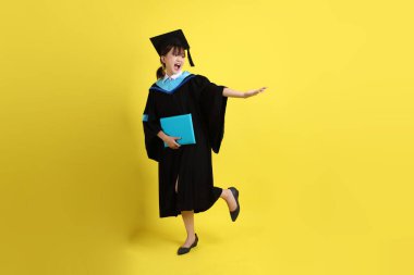 Mavi mezuniyet elbiseleri ve kepleri içinde kadın üniversite öğrencisi ve sarı arka planda izole edilmiş işaret eden bir jestle diplomaları var. Gen Z, yeni mezun, eğitim, ilk iş