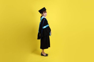 Mavi mezuniyet elbiseleri ve kepleri içinde, elinde diplomaları olan, sarı arka planda ayakta duran ve gülümseyen bir üniversite öğrencisi. Gen Z, yeni mezun, eğitim, ilk iş