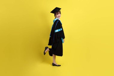 Mavi mezuniyet elbiseleri ve kepleri içinde, elinde diplomaları olan, sarı arka planda ayakta duran ve gülümseyen bir üniversite öğrencisi. Gen Z, yeni mezun, eğitim, ilk iş