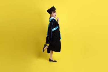 Mavi mezuniyet elbiseleri ve kepleri içinde kadın üniversite öğrencisi ve sarı arka planda izole edilmiş düşünce jestleriyle diplomaları var. Gen Z, yeni mezun, eğitim, ilk iş