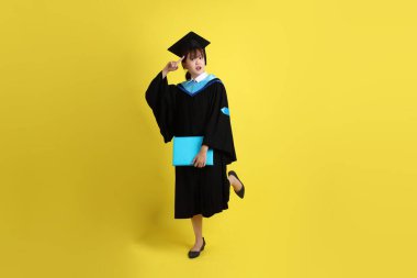 Mavi mezuniyet elbiseleri ve kepleri içinde kadın üniversite öğrencisi ve sarı arka planda izole edilmiş düşünce jestleriyle diplomaları var. Gen Z, yeni mezun, eğitim, ilk iş