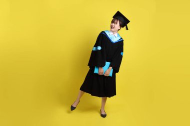 Mavi mezuniyet elbiseleri ve kepleri içinde, elinde diplomaları olan, sarı arka planda ayakta duran ve gülümseyen bir üniversite öğrencisi. Gen Z, yeni mezun, eğitim, ilk iş