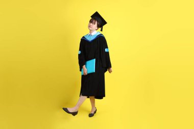 Mavi mezuniyet elbiseleri ve kepleri içinde kadın üniversite öğrencisi ve sarı arka planda izole bir şekilde yürüme jesti ile diplomaları var. Gen Z, yeni mezun, eğitim, ilk iş