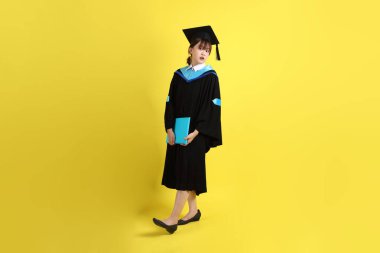 Mavi mezuniyet elbiseleri ve kepleri içinde, elinde diplomaları olan, sarı arka planda ayakta duran ve gülümseyen bir üniversite öğrencisi. Gen Z, yeni mezun, eğitim, ilk iş