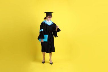 Mavi mezuniyet elbiseleri ve kepleri içinde, elinde diplomaları olan, heyecanlı, sarı arka planda izole bir şekilde kutlanan bir üniversite öğrencisi. Gen Z, yeni mezun, eğitim, ilk iş