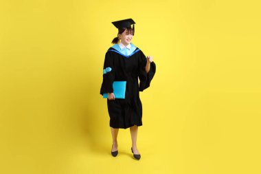 Mavi mezuniyet elbiseleri ve kepleri içinde, elinde diplomaları olan, heyecanlı, sarı arka planda izole bir şekilde kutlanan bir üniversite öğrencisi. Gen Z, yeni mezun, eğitim, ilk iş