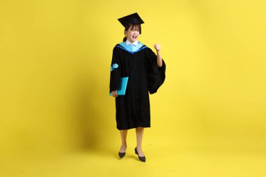 Mavi mezuniyet elbiseleri ve kepleri içinde, elinde diplomaları olan, heyecanlı, sarı arka planda izole bir şekilde kutlanan bir üniversite öğrencisi. Gen Z, yeni mezun, eğitim, ilk iş