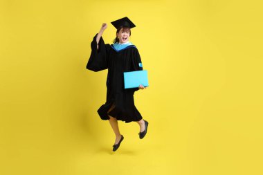 Mavi mezuniyet elbiseleri ve kepleri içinde kadın üniversite öğrencisi ve sarı arka planda izole bir şekilde atlama jesti ile diplomaları var. Gen Z, yeni mezun, eğitim, ilk iş