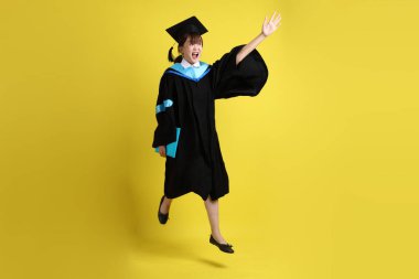 Mavi mezuniyet elbiseleri ve kepleri içinde kadın üniversite öğrencisi ve sarı arka planda izole bir şekilde atlama jesti ile diplomaları var. Gen Z, yeni mezun, eğitim, ilk iş