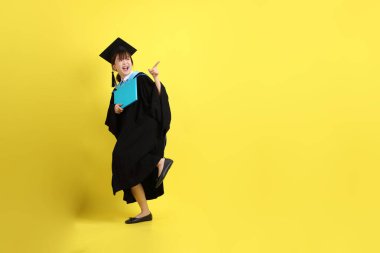 Mavi mezuniyet elbiseleri ve kepleri içinde kadın üniversite öğrencisi ve sarı arka planda izole edilmiş işaret eden bir jestle diplomaları var. Gen Z, yeni mezun, eğitim, ilk iş