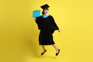 Mavi mezuniyet elbiseleri ve kepleri içinde kadın üniversite öğrencisi ve sarı arka planda izole bir şekilde atlama jesti ile diplomaları var. Gen Z, yeni mezun, eğitim, ilk iş