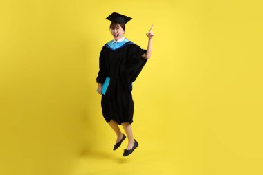 Mavi mezuniyet elbiseleri ve kepleri içinde kadın üniversite öğrencisi ve sarı arka planda izole bir şekilde atlama jesti ile diplomaları var. Gen Z, yeni mezun, eğitim, ilk iş