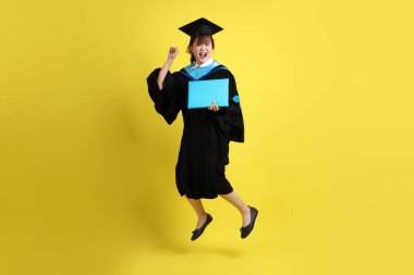 Mavi mezuniyet elbiseleri ve kepleri içinde kadın üniversite öğrencisi ve sarı arka planda izole bir şekilde atlama jesti ile diplomaları var. Gen Z, yeni mezun, eğitim, ilk iş