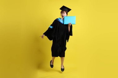 Mavi mezuniyet elbiseleri ve kepleri içinde kadın üniversite öğrencisi ve sarı arka planda izole bir şekilde atlama jesti ile diplomaları var. Gen Z, yeni mezun, eğitim, ilk iş