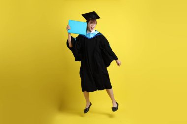 Mavi mezuniyet elbiseleri ve kepleri içinde kadın üniversite öğrencisi ve sarı arka planda izole bir şekilde atlama jesti ile diplomaları var. Gen Z, yeni mezun, eğitim, ilk iş