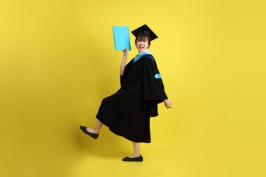 Mavi mezuniyet elbiseleri ve kepleri içinde kadın üniversite öğrencisi ve sarı arka planda izole bir şekilde atlama jesti ile diplomaları var. Gen Z, yeni mezun, eğitim, ilk iş