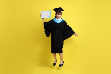 Mavi mezuniyet elbiseleri ve kepleri içinde kadın üniversite öğrencisi ve sarı arka planda izole bir şekilde atlama jesti ile diplomaları var. Gen Z, yeni mezun, eğitim, ilk iş