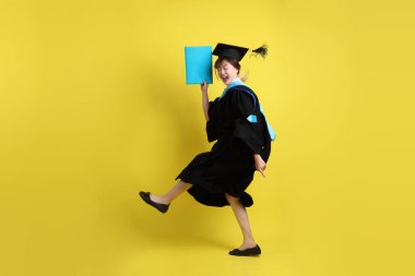 Mavi mezuniyet elbiseleri ve kepleri içinde kadın üniversite öğrencisi ve sarı arka planda izole bir şekilde atlama jesti ile diplomaları var. Gen Z, yeni mezun, eğitim, ilk iş