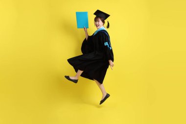 Mavi mezuniyet elbiseleri ve kepleri içinde kadın üniversite öğrencisi ve sarı arka planda izole bir şekilde atlama jesti ile diplomaları var. Gen Z, yeni mezun, eğitim, ilk iş