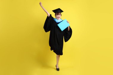 Mavi mezuniyet elbiseleri ve kepleri içinde, elinde diplomaları olan, heyecanlı, sarı arka planda izole bir şekilde kutlanan bir üniversite öğrencisi. Gen Z, yeni mezun, eğitim, ilk iş