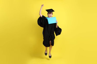 Mavi mezuniyet elbiseleri ve kepleri içinde kadın üniversite öğrencisi ve sarı arka planda izole bir şekilde atlama jesti ile diplomaları var. Gen Z, yeni mezun, eğitim, ilk iş