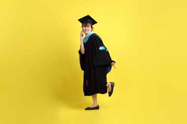 Mavi mezuniyet elbisesi ve şapkalı, sarı arka planda izole cep telefonu bulunduran bir kız öğrenci. Gen Z, yeni mezun, eğitim, ilk iş