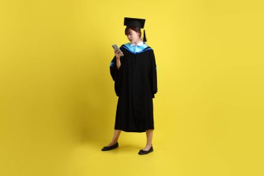 Mavi mezuniyet elbisesi ve şapkalı, sarı arka planda izole cep telefonu bulunduran bir kız öğrenci. Gen Z, yeni mezun, eğitim, ilk iş